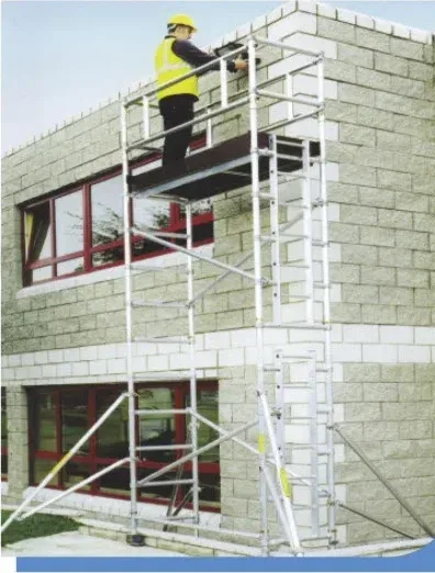 Worker in a yellow safety vest standing on an aluminum scaffold tower beside a stone-block building