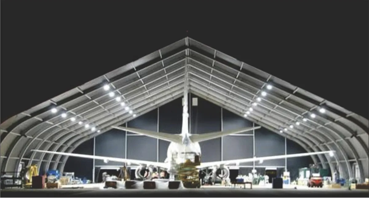 Large curved clear-span hangar tent illuminated at night with an aircraft parked inside